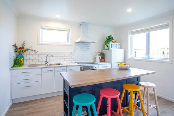 Modern kitchen with white cabinets, colorful stools, fruit bowl, and plants on a wooden island table.