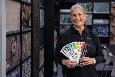 Woman holding color samples, smiling in store with wallpaper displayed in background.