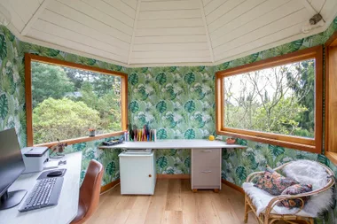 Tropical-themed home office with large windows, leafy wallpaper, desk, chair, and computer; view of greenery outside.