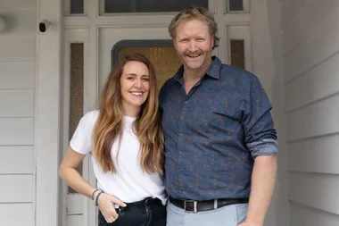 Man and woman smiling and standing together at a front door, casual attire.