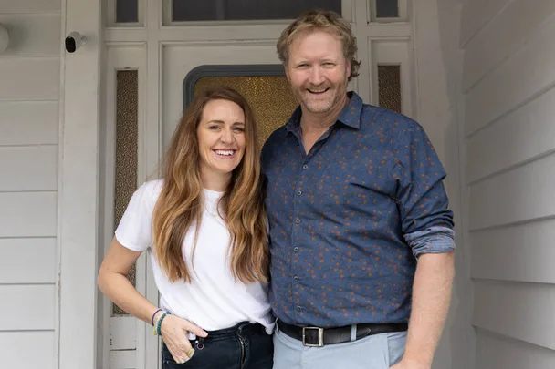 Man and woman smiling and standing together at a front door, casual attire.