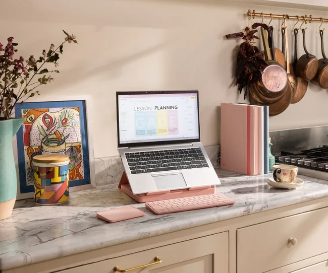 A laptop on a kitchen counter with lesson planning screen, a keyboard, a pink notebook, and decorative items.