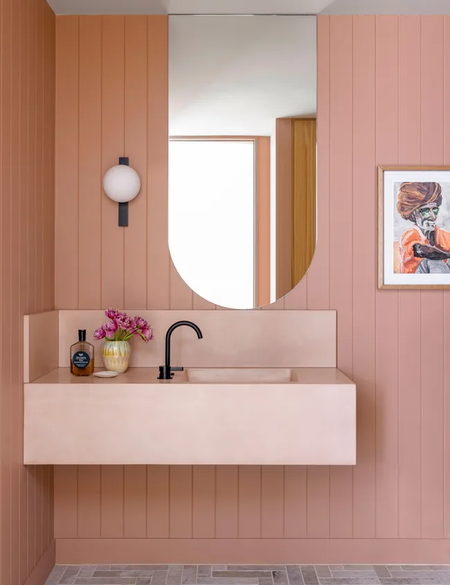 Peach bathroom with wall-mounted sink, black faucet, round mirror, wall sconce, purple flowers, and a framed painting.