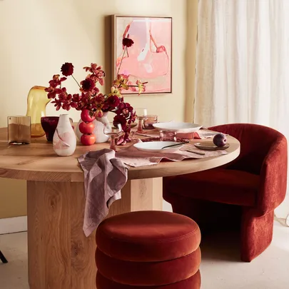Round wooden table set with red decor, flowers, and velvet chairs in a warmly lit dining room.
