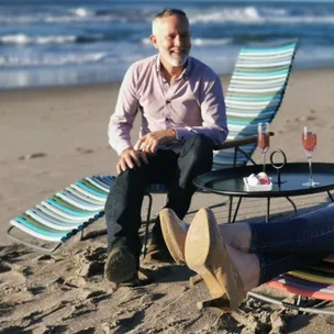 Man sitting on a beach chair with drinks on a table, ocean in the background.