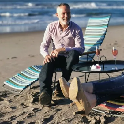 Man sitting on a beach chair with drinks on a table, ocean in the background.
