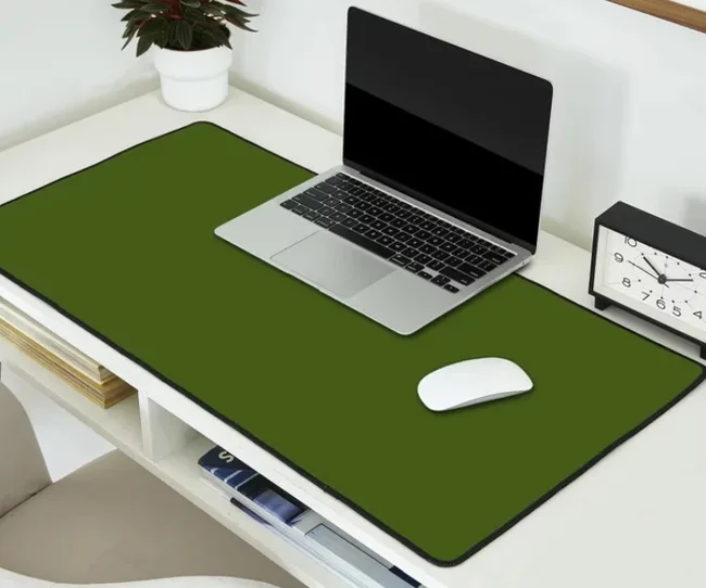 Laptop on a large green desk mat with a mouse and clock nearby, set on a white desk with a potted plant.