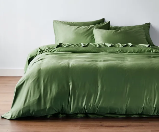 Green satin bed set with two pillows on a wooden floor background.