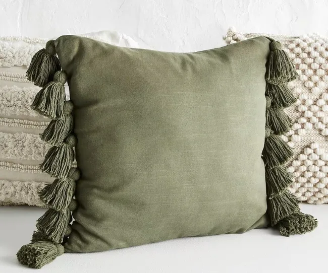 Green throw pillow with tassels on sides, placed on a textured cream-colored pillow background.