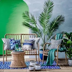 Outdoor seating area with rattan furniture, blue patterned cushions, palm plants, and green wall background.