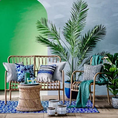 Outdoor seating area with rattan furniture, blue patterned cushions, palm plants, and green wall background.