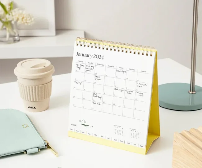 Table calendar displaying January 2024 on a desk with a coffee cup, lamp, and a mint pouch.