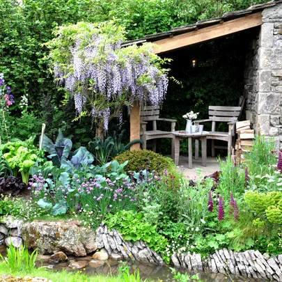Rustic garden with blooming flowers, lush greenery, and a stone alcove with two wooden chairs under wisteria.