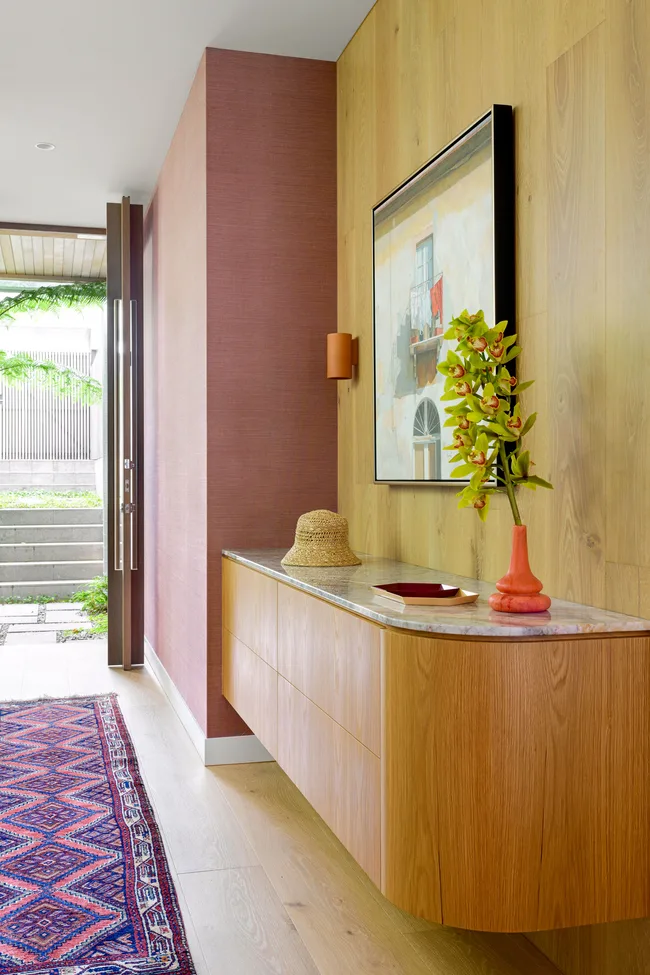 Modern hallway with wood console, orchid vase, straw hat, and vibrant rug, leading to an open glass door.