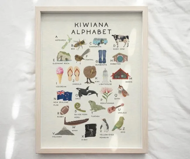 Framed "Kiwiana Alphabet" print with illustrated New Zealand icons for each letter, like kiwi, gumboots, and pohutukawa.