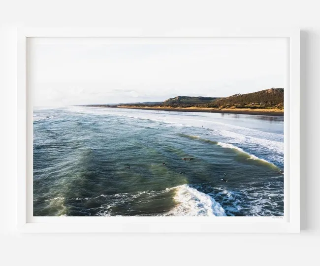 Framed photo of surfers in the ocean with waves and a distant sandy coastline under a clear sky.