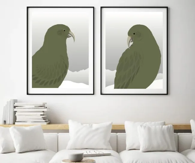 Two framed prints of green parrots on the wall above a white sofa with cushions and stacked books.