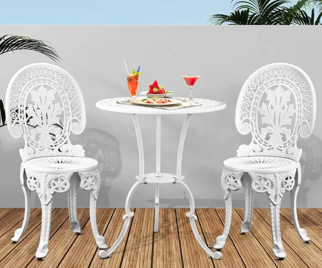 White ornate patio set with two chairs and a table, featuring drinks and a dessert, on a wooden deck with palm plants.