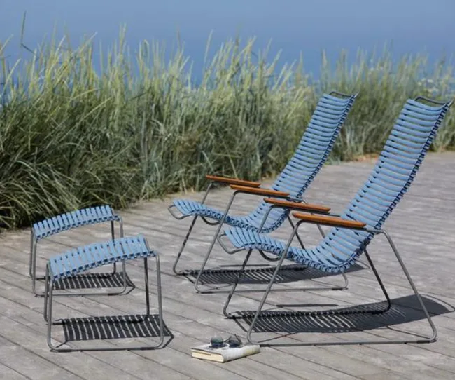 Blue outdoor chairs and footstools on a wooden deck, with grasses and ocean view in the background.