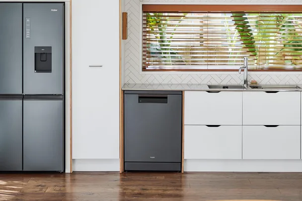 Modern kitchen with Haier fridge, dishwasher, white cabinets, wooden floor, and large window with blinds.