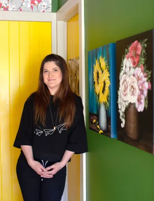 A woman in a black shirt stands in a colorful room with floral paintings on the wall.
