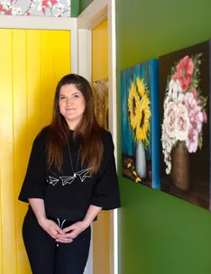 A woman in a black shirt stands in a colorful room with floral paintings on the wall.
