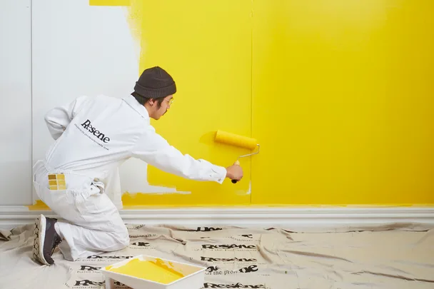 Painter in Resene overalls applying yellow paint with a roller on a wall, using a paint tray on a drop cloth.