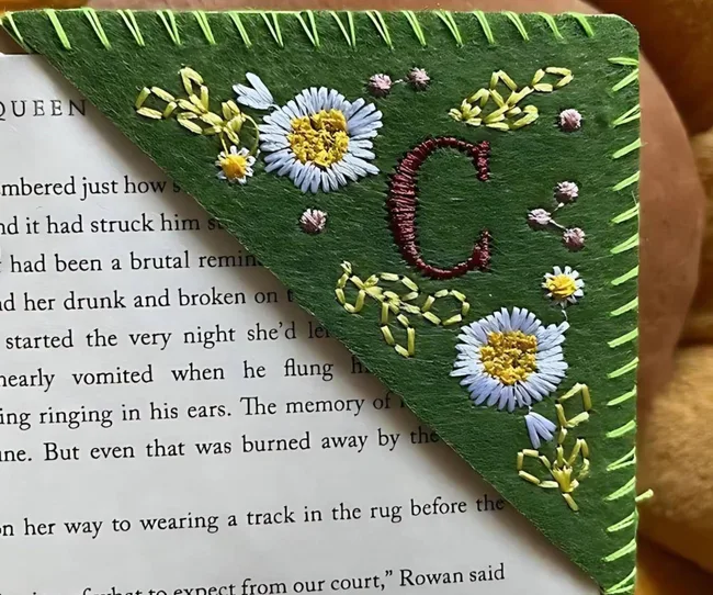 A green embroidered bookmark with the letter C and flowers on it, over the corner of a book