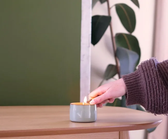 Hand lighting a small candle on a wooden table with green wall and plant in the background.