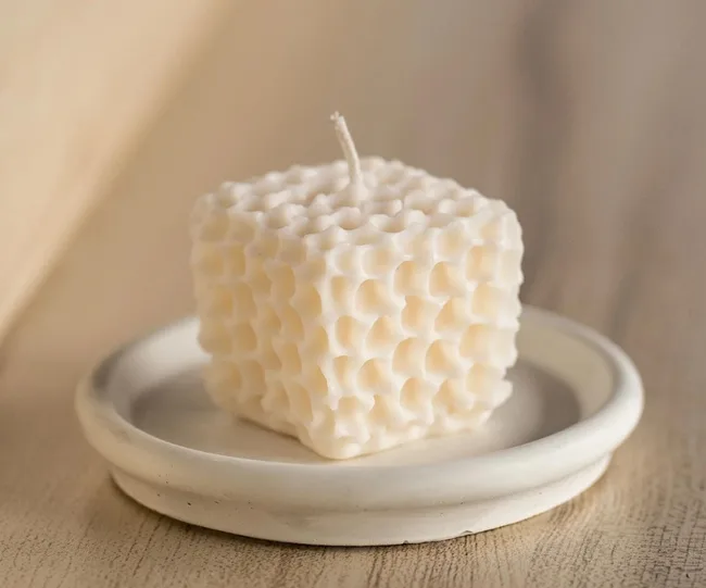Cream-colored textured cube candle on a white ceramic dish, placed on a wooden surface.