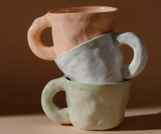 Three handmade ceramic mugs stacked; colors are peach, white, and light green on a neutral background.