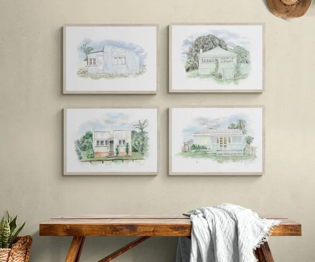 Four framed watercolor house paintings on a beige wall above a wooden bench with a light gray throw.