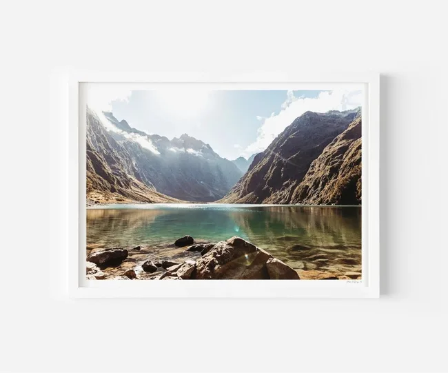 Framed photograph of sunny mountain landscape with a calm lake, clear sky, and rocky foreground.