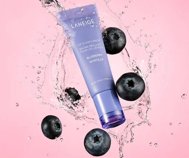 A purple Laneige lip balm surrounded by blueberries against a pink background