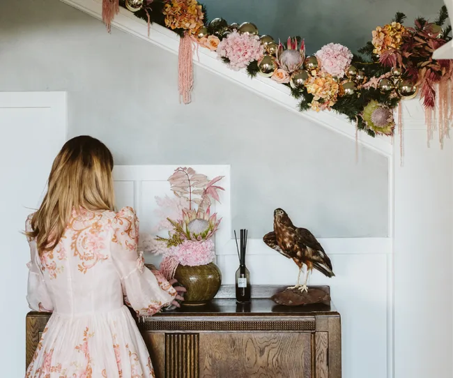 A woman in a floral dress arranging pink flowers on a cabinet under a festive garland near a bird sculpture.