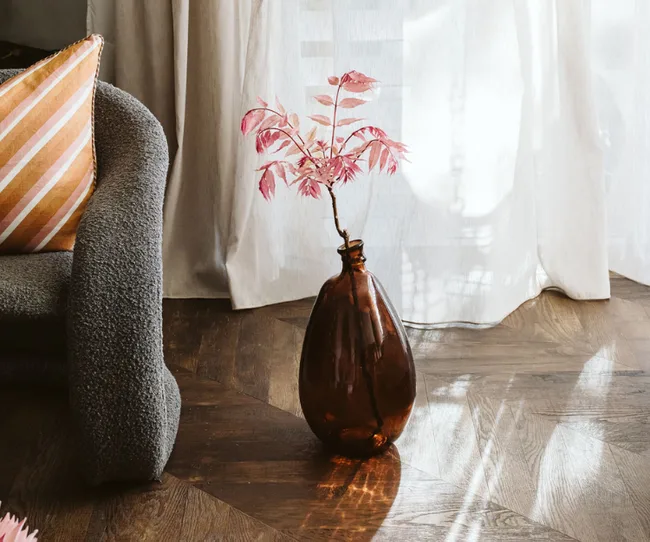 A brown glass vase with pink leaves on a wooden floor, next to a grey chair and striped pillow, with sunlight streaming in.
