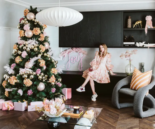 Woman sitting by a decorated Christmas tree with pink and gold ornaments, beside a neon "Joy" sign in a stylish room.