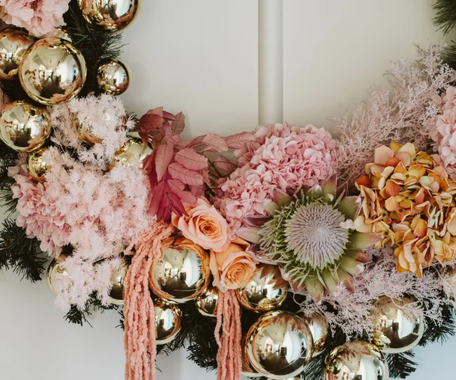 Festive wreath with pink and peach flowers, reflective gold ornaments, and greenery on a white background.