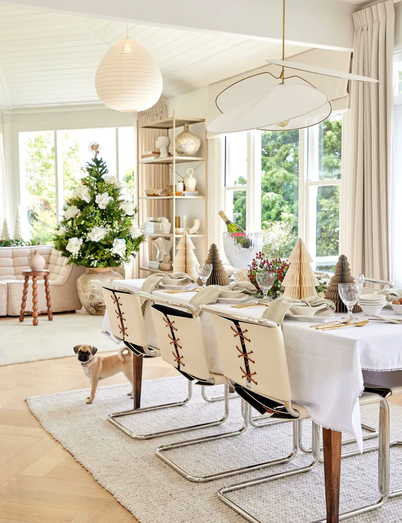 Festive dining room with table set for holiday, modern decor, Christmas tree, and a small dog on the floor.