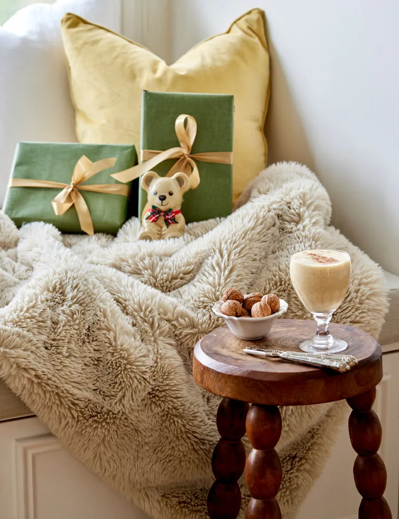 Cozy corner with wrapped gifts, a teddy bear, a fluffy blanket, a small table with nuts, and a glass of creamy drink.