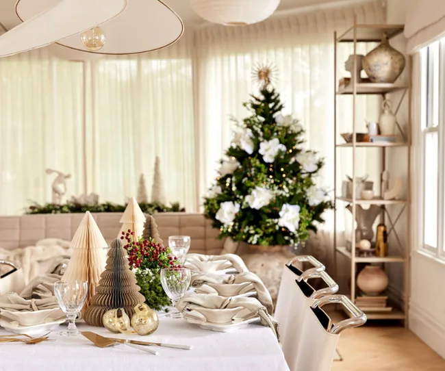 Festive dining table with decorations and a Christmas tree with white flowers in a bright room.