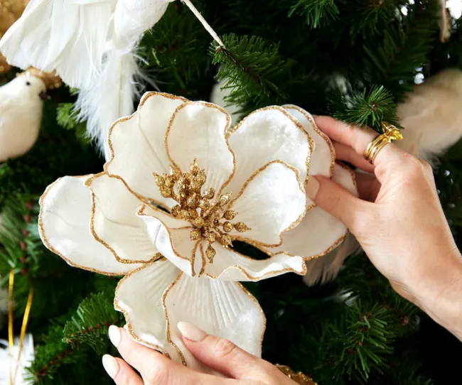 Hands placing a white and gold floral decoration on a Christmas tree.