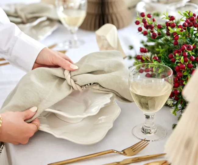 Hands arranging a knotted napkin on stacked dishware with wine glass and red berry centerpiece on a dining table.