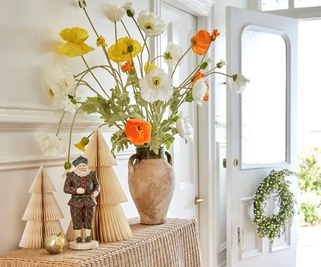 Decorative flowers in a vase, paper trees, and a festive figurine on a wicker table by an open door with a wreath.