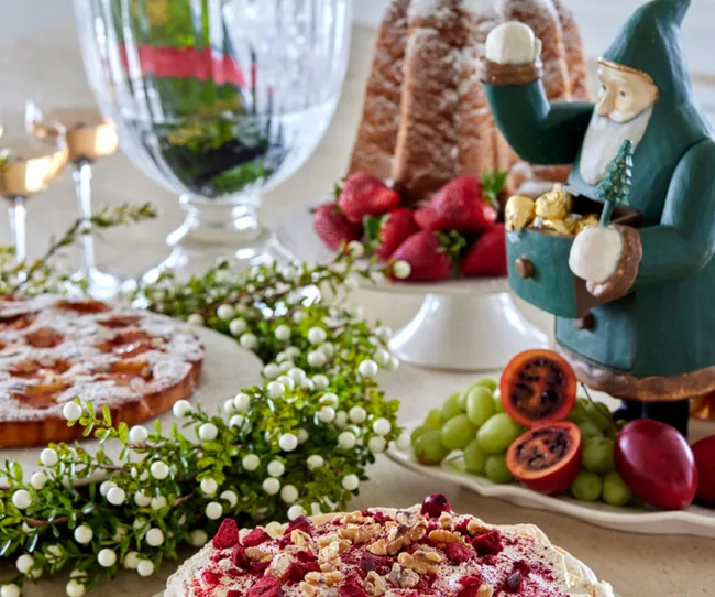 Festive table setting with sweets, fruits, a green Santa figurine, greenery, and candles.