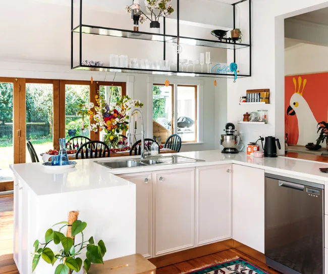 Modern kitchen with white cabinets, floral centerpiece, hanging shelves, and bird artwork. Bright and airy.
