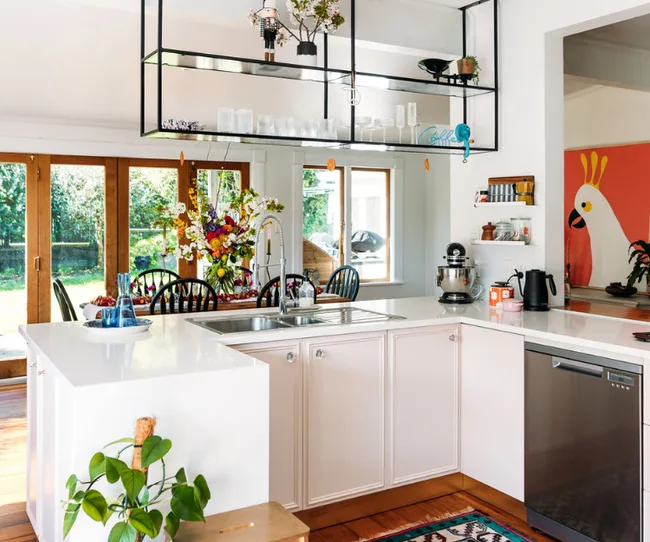 Modern kitchen with white cabinets, floral centerpiece, hanging shelves, and bird artwork. Bright and airy.