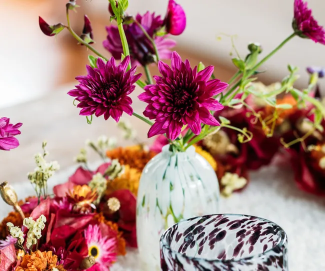 Purple flowers in a white vase surrounded by colorful petals on a table.