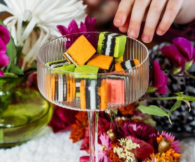 A hand reaching for colorful striped candies in a glass, surrounded by vibrant flowers.
