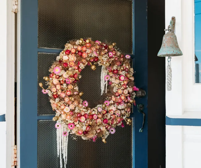 A vibrant dried flower wreath with pink and gold hues hangs on a blue door next to a rustic metal bell.
